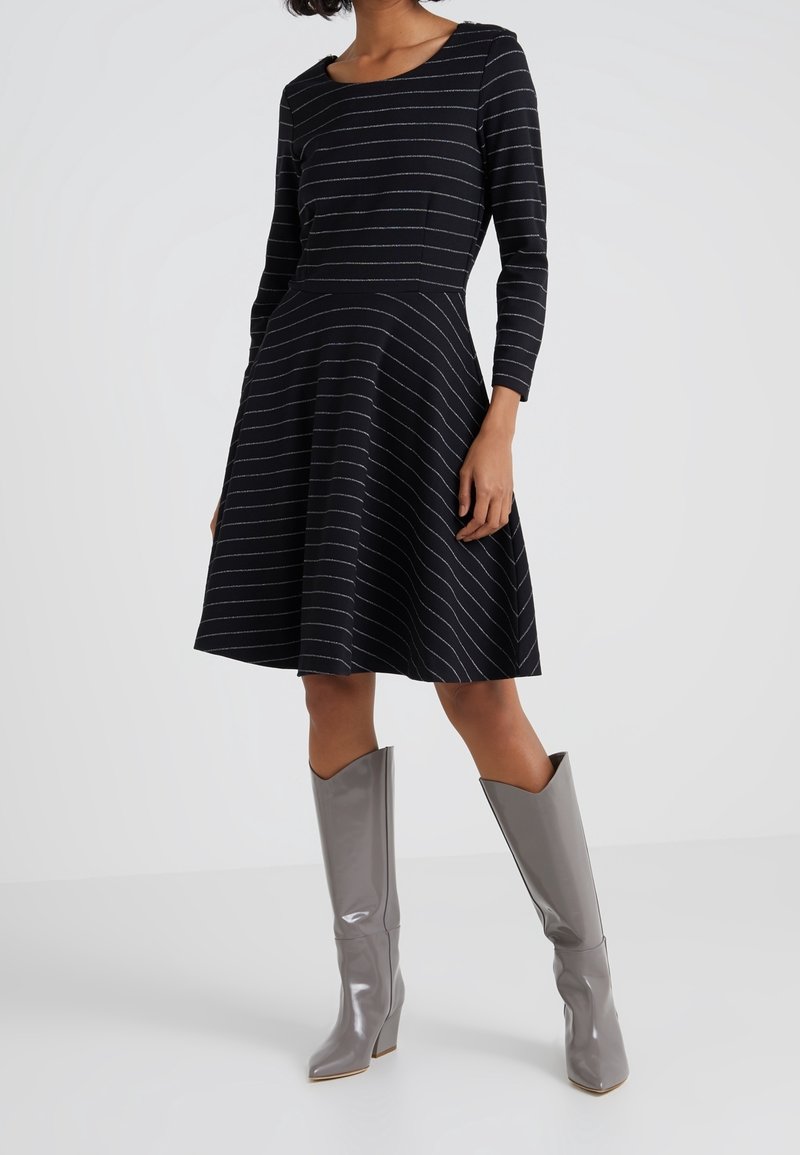 Femme portant une robe noire à manches longues, longueur genoux, avec de fines rayures blanches horizontales et des bottes grises brillantes montantes jusqu'aux genoux.