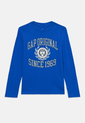 Kék, hosszú ujjú póló, kerek nyakkivágással. Az elején szürkével nyomtatva van a "GAP ORIGINAL SINCE 1969" felirat, valamint középen egy kör alakú logó található.