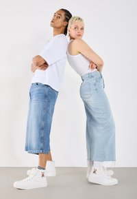 Deux individus se tenant dos à dos, portant des jeans en denim léger et ample, des tops blancs et des baskets blanches. Une des baskets a un patch décoratif.