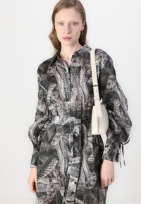 Vestito a motivo bianco e nero con design a bottoni, maniche lunghe e vita con cintura. Tiene una piccola borsa color crema.