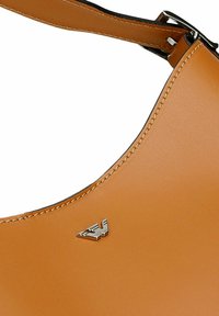 Lichte leren handtas met zilveren gesp en klein zilveren adelaarlogo op een glad oppervlak, met gedetailleerde stiksels langs de rand.