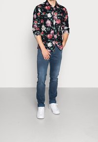 Blommig skjorta med knappar i svart med rosa och vita blommor, tillsammans med blå jeans och vita sneakers. Avslappnad passform, med vikta ärmslut synliga.