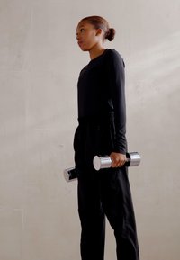 Black athletic outfit with long sleeves, holding metal dumbbells. The background is a smooth, light-colored wall.