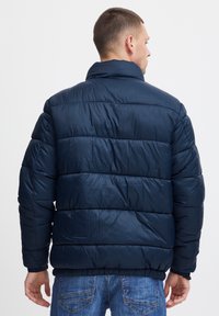 Marineblå pufferjakke med høj krave og quiltet design, der har en glat, skinnende tekstur og elastiske manchetter, båret over blå jeans.