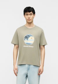 Young man wearing light olive green T-shirt with a beach scene and "Jack & Jones" text, paired with light blue jeans.