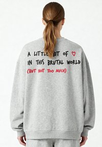 Felpa grigia oversize con maniche lunghe. Sul retro presenta un testo nero e rosso: "UN PO' DI ♥ IN QUESTO MONDO BRUTALE (MA NON TROPPO)."