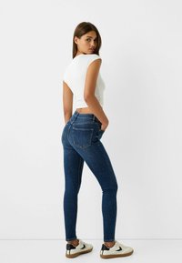 Tajta jeans i mörkblå denim med hög midja, bakfickor och en slät yta. Kombinerat med en åtsittande vit topp och sneakers.