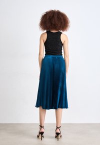 Top nero senza maniche abbinato a una gonna midi plissettata color teal e décolleté neri con tacco a spillo. La gonna presenta pieghe verticali e una texture liscia.