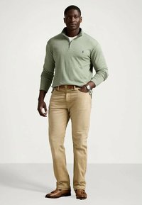 Άνδρας που φοράει ανοιχτό πράσινο πουλόβερ με φερμουάρ μέχρι τον ώμο, μπεζ παντελόνι, καφέ ζώνη και καφέ loafers με τασέλες, στέκεται με το ένα χέρι στην τσέπη.