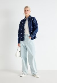 Jaqueta de denim com bordados florais, top cinza, calças de ganga largas azuis claras e mala branca. Modelo está a usar ténis brancos.