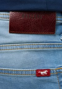 Niebieskie dżinsy z teksturowaną brązową łatką z napisem "MUSTANG" oraz czerwoną metką z białym logo konia w pobliżu kieszeni.