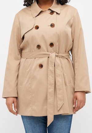 Personne portant un trench-coat beige à double boutonnage avec des boutons foncés et une ceinture nouée à la taille, associé à un jean bleu.