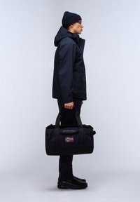 Námornícká duffle taška s černými akcenty, se znakem norské vlajky, nesena osobou v odpovídajícím námořnickém kabátu a beanie.