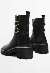 Bottes noires mi-mollet avec des talons épais, des semelles à crampons, deux boucles dorées sur le côté extérieur et une fermeture éclair sur le côté intérieur de chaque botte.