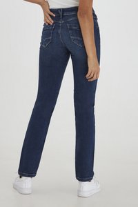 Vaqueros de mezclilla en azul oscuro con un corte ajustado, cintura alta y dos bolsillos traseros. Combinados con zapatillas blancas; textura suave y diseño clásico.