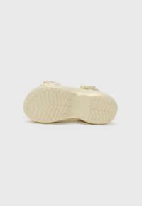 Sandale avec semelle beige texturée tournée vers le haut, montrant le motif de la semelle et une petite bride arrière avec des éléments décoratifs.