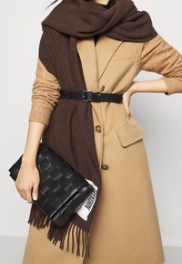 Połączenie beżowego wełnianego płaszcza z brązowym szalem i czarnym plecionym kopertówką. Płaszcz ma dopasowany krój i guziki; szal ma frędzle.