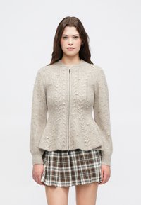 Jovem mulher com cabelo longo e escuro, vestindo um suéter de malha bege com detalhes em pérola e uma saia plissada xadrez castanha, voltada para a frente.