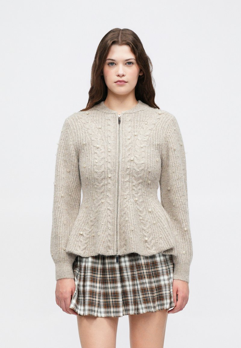 Jovem mulher com cabelo longo e escuro, vestindo um suéter de malha bege com detalhes em pérola e uma saia plissada xadrez castanha, voltada para a frente.