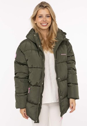Lächelnde Frau mit langen blonden Haaren trägt eine olivgrüne Steppjacke über einem weißen Hemd und weißen Hosen vor weißem Hintergrund.