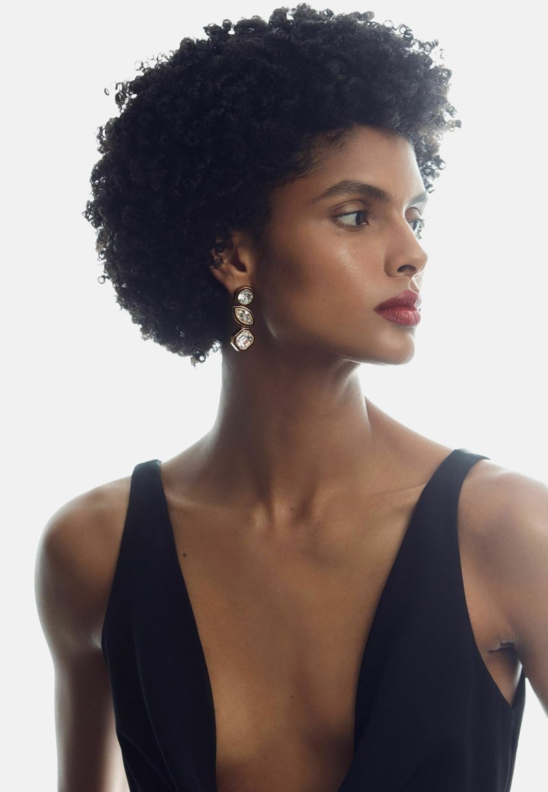 Žena s krátkými kudrnatými vlasy, která má na sobě černé šaty s hlubokým vykrojením do V a dlouhé geometrické náušnice, dívající se doprava na bílém pozadí.