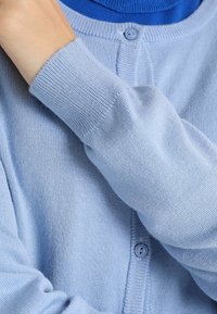 Jasnoniebieski kardigan wykonany z miękkiego materiału, z okrągłym dekoltem, długimi rękawami i zapięciem na guziki z prążkowaną teksturą.