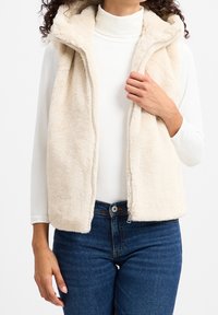 Gilet in pile beige chiaro, con colletto alto e zip frontale. Tessuto morbido e vestibilità casual, abbinato a un top bianco a maniche lunghe.