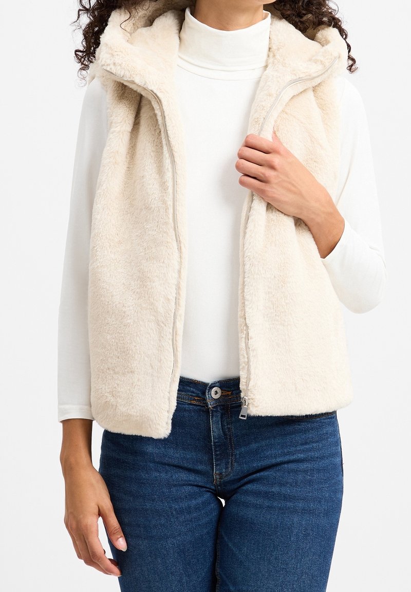 Gilet in pile beige chiaro, con colletto alto e zip frontale. Tessuto morbido e vestibilità casual, abbinato a un top bianco a maniche lunghe.