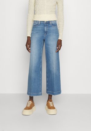 Jean denim bleu clair à jambes larges avec un ourlet brut, associé à des baskets plateformes marron. Le haut est un chemisier crème texturé à manches longues.