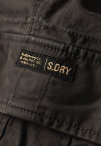 Tissu noir avec une texture subtile, comportant une étiquette sur laquelle est inscrit "S.DRY" en lettres dorées. Les coutures surpiquées sont visibles, indiquant la durabilité.