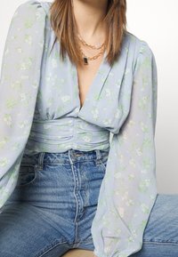 Ljusblå blommig blus med långa ärmar och rynkad midja, i kombination med högmidjade jeans i denim. Blommönstret har gröna inslag.