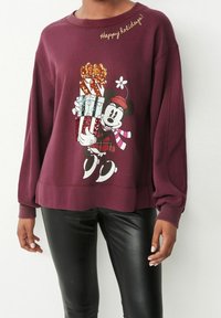 Sweatshirt bordeaux avec un graphique coloré et pailleté d'un personnage tenant des cadeaux, avec "Joyeuses fêtes !" en texte doré. Tissu doux, coupe décontractée.