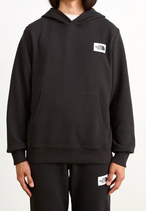 Osoba ubrana w czarną bluzę z kapturem The North Face oraz pasujące spodnie z białymi naszywkami z logo, stojąca na jednolitym tle.