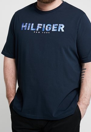 Mand iført en mørkeblå t-shirt med teksten "HILFIGER NEW YORK" i store blå og hvide bogstaver hen over brystet.