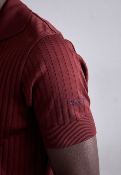 Polo shirt à manches courtes rouge à côtes avec col, présentant un logo brodé bleu sur la manche gauche et un tissu texturé.