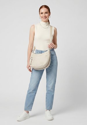 Vrouw staat, glimlacht, draagt een mouwloos crème topje, lichtblauwe spijkerbroek, witte sneakers en draagt een beige schoudertas.