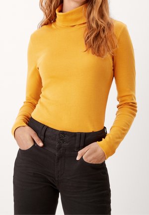Pull à col roulé à manches longues jaune moutarde associé à un pantalon taille haute noir avec fermeture à deux boutons et poches.