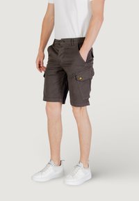 Pantaloni cargo marrone scuro con una tasca laterale, chiusura con bottone e passanti per cintura, abbinati a sneaker bianche e una t-shirt bianca semplice.