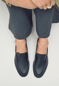 Mustad musta nahast loaferid, millel on õmblustäike, ümar nina ja sile tekstuur. Kantud hallide lühikeste pükste juurde.