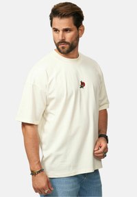 Cremfarbener Baumwoll-T-Shirt mit lockerer Passform, kurzen Ärmeln und einem kleinen gestickten Design auf der Brust. Kombiniert mit blauen Jeans.