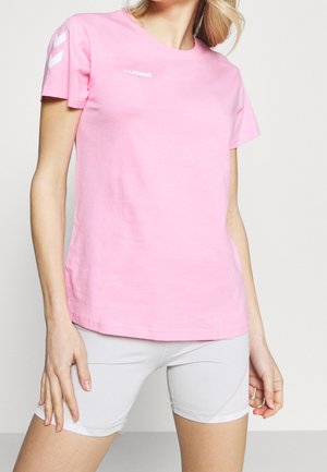 Vrouw draagt lichtroze hummel T-shirt met korte mouwen en witte aansluitende shorts, staand met ontspannen handen langs haar zij.