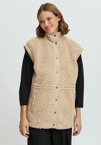 Beige, ärmellose Weste mit weichem Kunstfell-Textur, Druckknöpfen, hohem Kragen und seitlichen Taschen. Glatter, kontrastierender Stoff auf den Schultern.