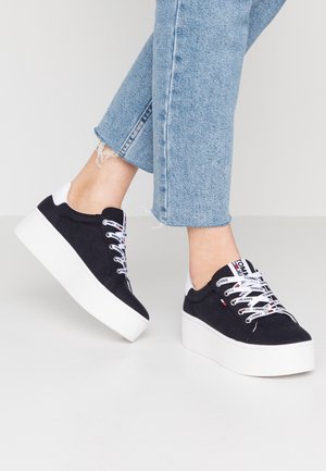 Czarne sneakersy na platformie z białymi grubymi podeszwami, białe sznurowadła z logo, noszone z jasno-niebieskimi, krótkimi postrzępionymi jeansami.