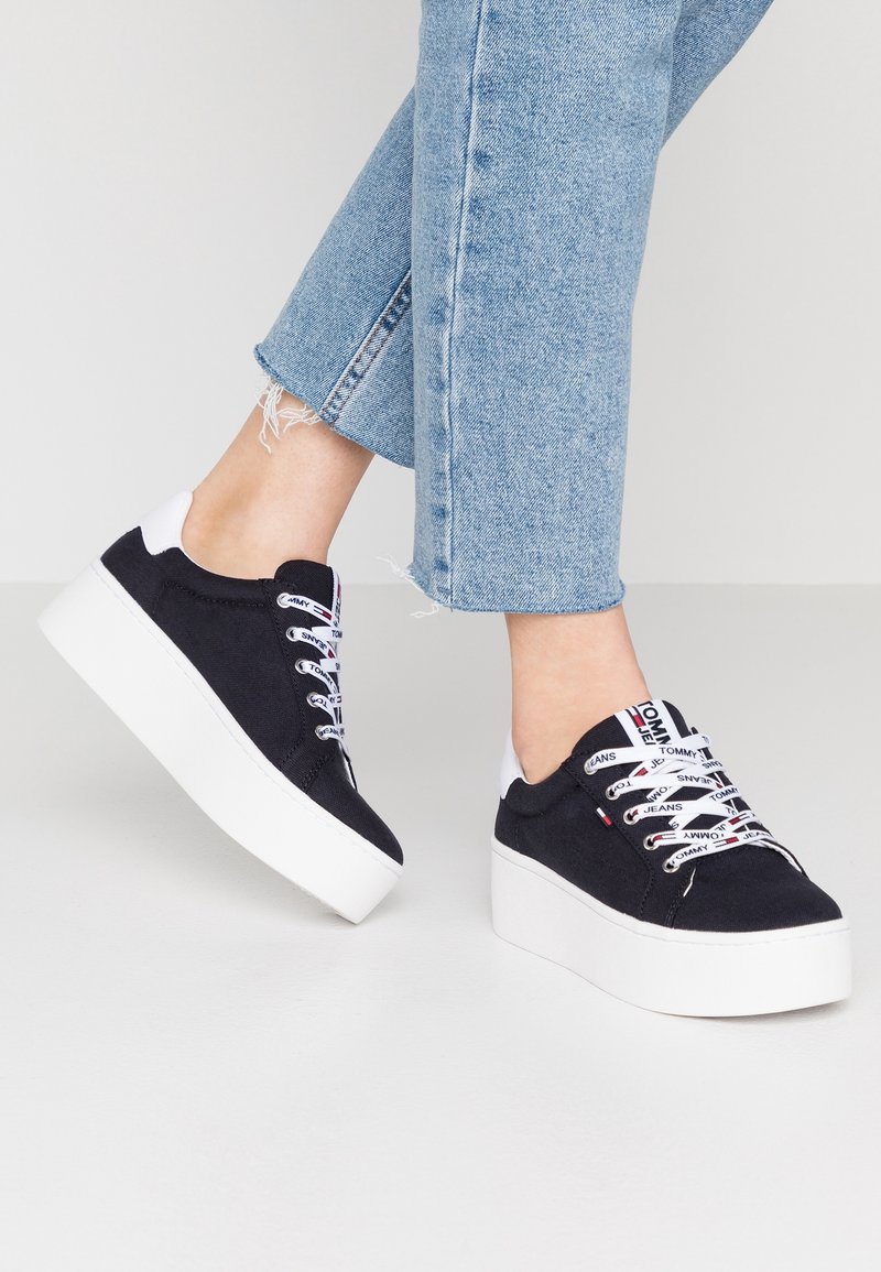 Czarne sneakersy na platformie z białymi grubymi podeszwami, białe sznurowadła z logo, noszone z jasno-niebieskimi, krótkimi postrzępionymi jeansami.