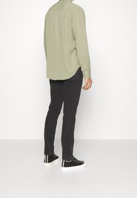 Camisa de manga larga verde claro para hombre, pantalones ajustados negros y zapatillas negras con suelas blancas, vistas desde la espalda.
