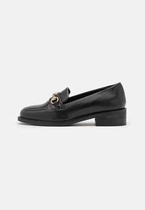 Mocassins en cuir noir avec un bout arrondi, un talon de hauteur moyenne et une boucle dorée décorative à l'avant. Texture lisse et design épuré.