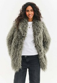 Kobieta o długich kręconych włosach ma na sobie oversize'owy szary futrzany płaszcz, białą koszulę, czarne jeansy i warstwowe naszyjniki, uśmiecha się do aparatu.