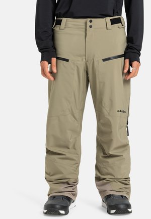 Pantalons de neige imperméables beiges avec poches zippées, taille ajustable et coupe ample ; poignets renforcés d'une teinte plus foncée. Bottes noires visibles.