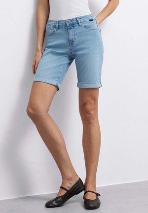 Persona che indossa pantaloncini di denim azzurro con orlo arrotolato, top bianco e scarpe Mary Jane nere, in piedi contro una parete bianca liscia.