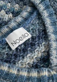 Blødt strikket stof i blå og hvide nuancer med en hvid label, der viser mærkenavnet "Noella", syet på.